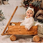 Aria participe au concours pour gagner de l'argent avec cette photo : baby, child, wagon, wooden_wagon, white_dress, bow_headband, winter_decor, pine_cones, christmas_tree, holiday_theme, smiling, portrait, indoor, cozy, decorations, cute, infant, festive, soft_lighting, warm_colors