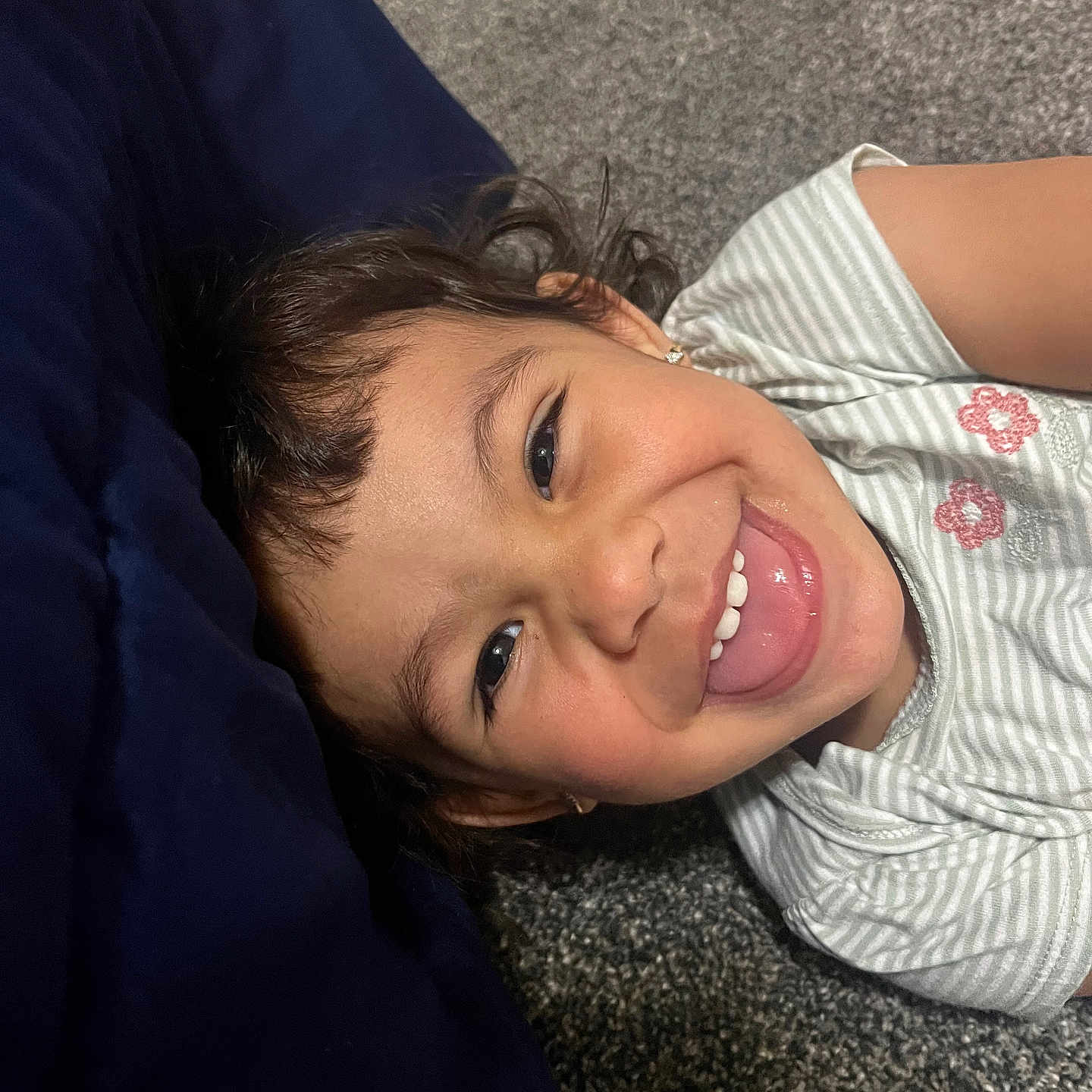 Allyson is registered to the contest to win money with this photo: child, smiling, tongue_out, striped_shirt, floral_embroidery, earrings, carpet, indoor, playful, happy, portrait, closeup, person, young, cute, casual_clothing, lying_down, headshot, face, fun