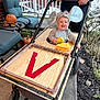 adult_hand, black_clothing, brick_wall, child, costume, fall_decor, happy, mouse_costume, outdoor_furniture, plants, porch, pumpkin, sidewalk, smiling, stroller, toddler, toy_cheese, wheel, wooden_panel, yellow_sponge