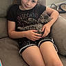 animal, black_tshirt, carpet, cat, child, couch, cute, girl, holding, home, indoor, kitten, light, pet, relaxing, seated, shorts, smile, window, young