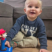 Nathanaël participe au concours pour gagner de l'argent avec cette photo : toddler, child, boy, smiling, indoor, toy, mario, sweater, pants, socks, wooden_floor, couch, living_room, blonde_hair, happy, playing, floor, casual_clothing, blue_wall, person