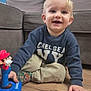 toddler, child, boy, smiling, indoor, toy, mario, sweater, pants, socks, wooden_floor, couch, living_room, blonde_hair, happy, playing, floor, casual_clothing, blue_wall, person