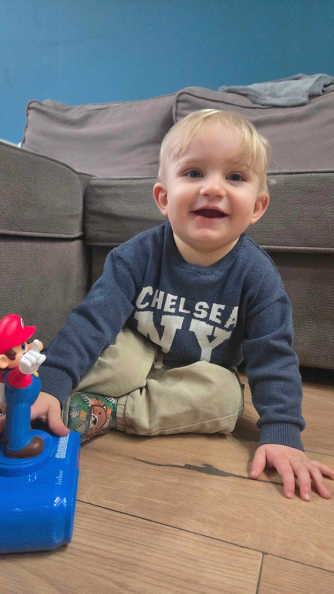 Nathanaël participe au concours pour gagner de l'argent avec cette photo : toddler, child, boy, smiling, indoor, toy, mario, sweater, pants, socks, wooden_floor, couch, living_room, blonde_hair, happy, playing, floor, casual_clothing, blue_wall, person