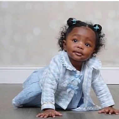 Sarai is registered to the contest to win money with this photo: toddler, child, baby, curly_hair, pigtails, blue_bows, crawling, floor, light_blue_clothing, ruffled_collar, long_sleeves, indoor, neutral_background, cute, portrait, young_child, person, happy, curious, small_hands