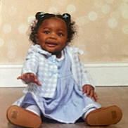 Sarai is registered to the contest to win money with this photo: baby, baseboard, blue_bows, blurry, child, curly_hair, cute, dress, floor, footwear, indoor, jacket, person, portrait, reaching_hand, sitting, smiling, toddler, wall, young_child