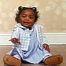 baby, baseboard, blue_bows, blurry, child, curly_hair, cute, dress, floor, footwear, indoor, jacket, person, portrait, reaching_hand, sitting, smiling, toddler, wall, young_child