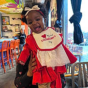 Sarai joined the competition — help win amazing prizes! baby, child, smiling, red_clothing, bib, indoor, restaurant, colorful, curtains, table, chair, person_holding_baby, woman, window, flower_pattern, headband, hair_bow, happy, portrait, celebration