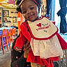 baby, child, infant, red_clothing, bib, bow, curly_hair, smiling, indoor, person_holding_child, colorful_background, orange_chairs, tablecloth, festive, cute, portrait, happy, toddler, headband, celebration