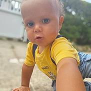 Milow a rejoint le concours — aidez-le/la à gagner de superbes lots ! baby, blurred_background, casual_clothing, child, closeup, crawling, curious, cute, daylight, denim_overalls, expression, face, grass, hand, outdoor, person, portrait, toddler, yellow_shirt, young_child