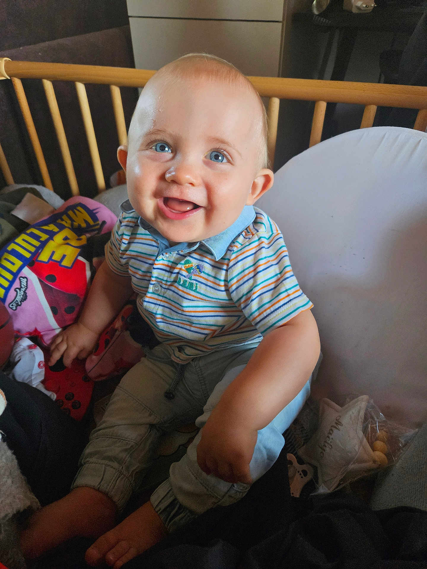 Maëlyo participe au concours pour gagner de l'argent avec cette photo : baby, child, smile, blue_eyes, striped_shirt, polo_shirt, crib, wooden_crib, toy, blanket, indoor, cute, happy, infant, person, sitting, footwear_free, playful, closeup, portrait