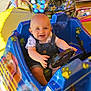 baby, child, amusement_ride, kiddie_ride, steering_wheel, blue_car, cartoon_characters, outdoor, daylight, smiling, happy, playful, shorts, overalls, seat, toy_car, bright_colors, sunlight, face, eyes