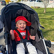 Luca joined the competition — help win amazing prizes! baby, child, stroller, red_cap, red_shirt, nike_logo, smiling, happy, outdoor, sunny, suburban, grass, tree, house, sidewalk, car, seatbelt, person, infant, casual_clothing