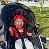 baby, child, stroller, red_cap, red_shirt, nike_logo, smiling, happy, outdoor, sunny, suburban, grass, tree, house, sidewalk, car, seatbelt, person, infant, casual_clothing