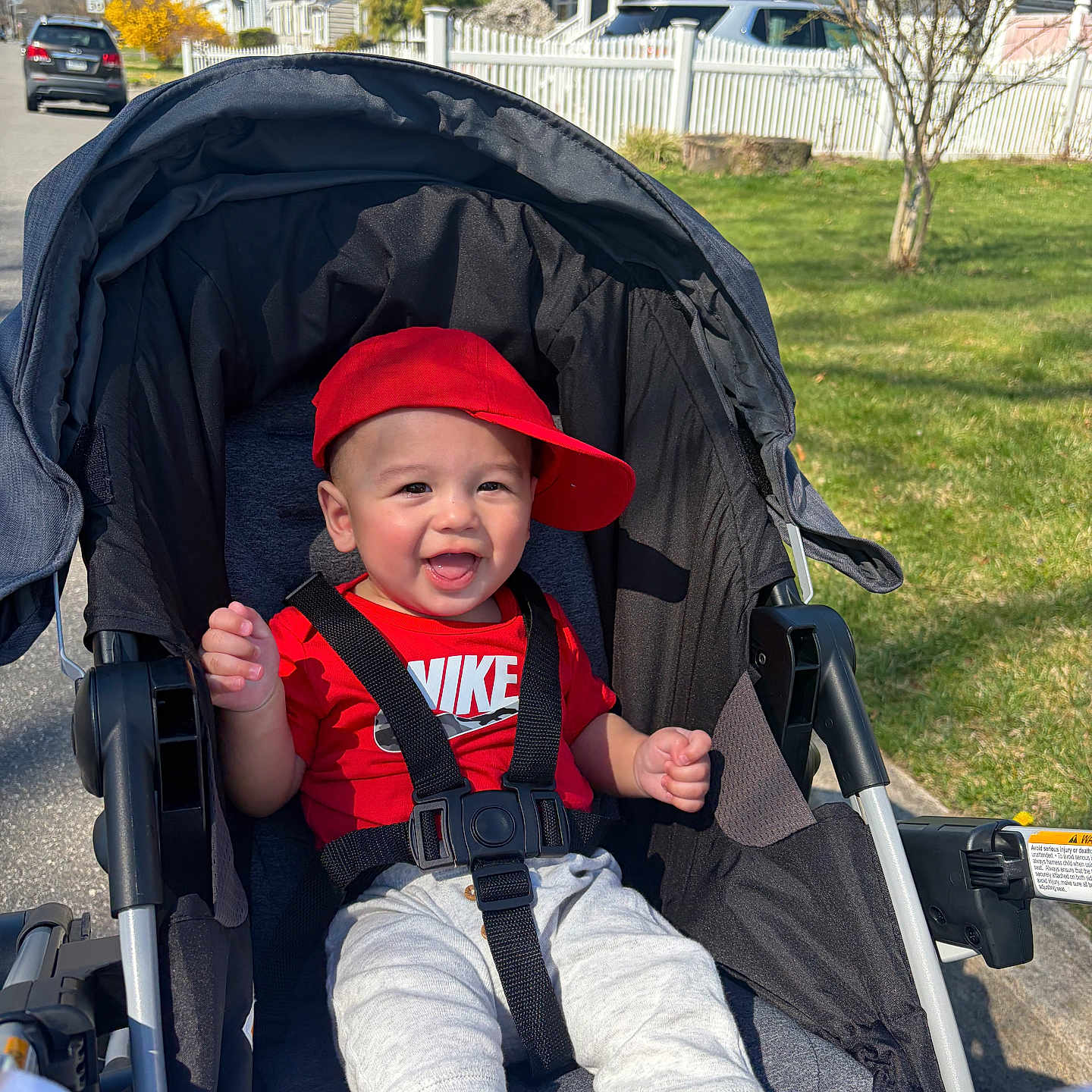 Luca joined the competition — help win amazing prizes! baby, child, stroller, red_cap, red_shirt, nike_logo, smiling, happy, outdoor, sunny, suburban, grass, tree, house, sidewalk, car, seatbelt, person, infant, casual_clothing
