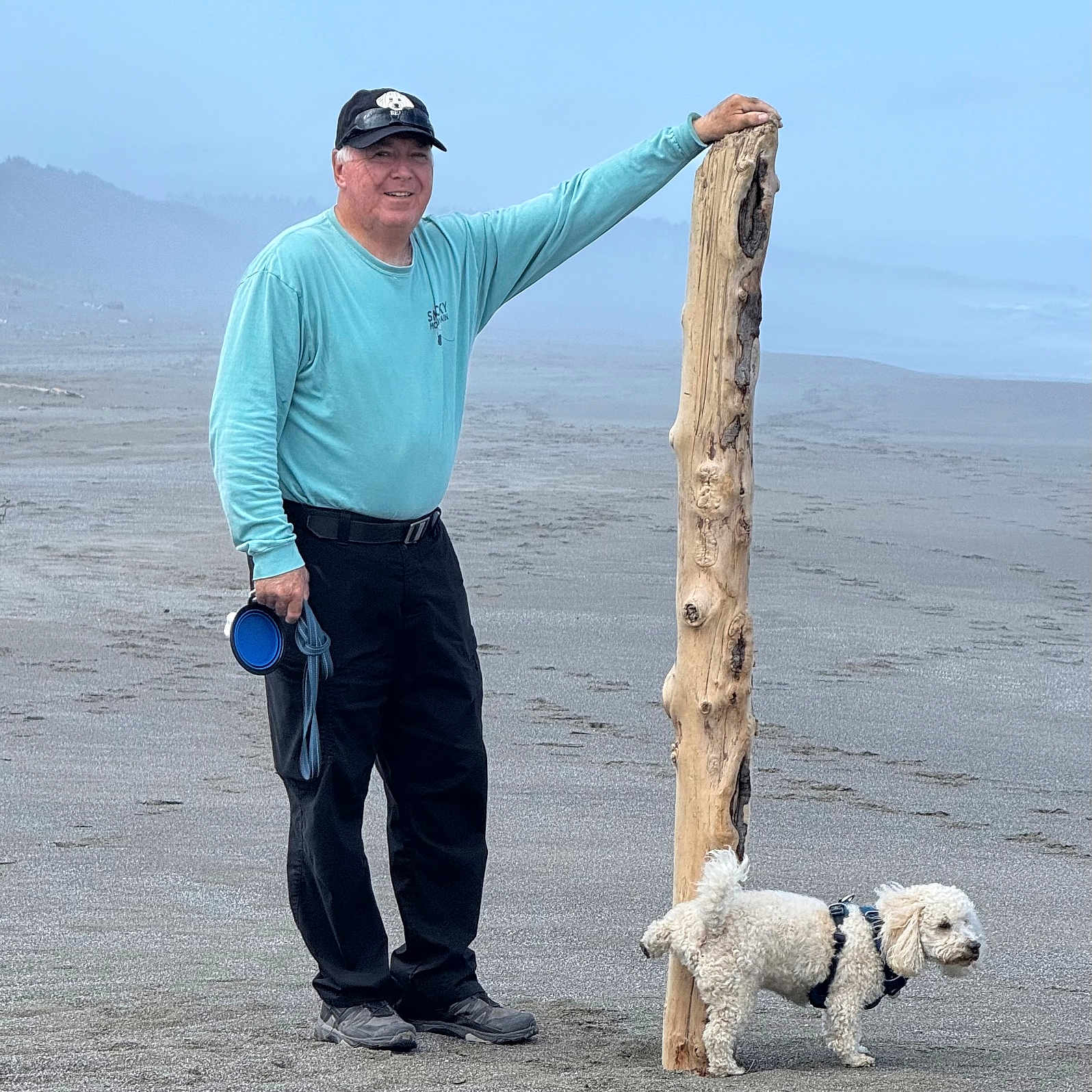 Ready is registered to the contest to win money with this photo: beach, canine, casual_clothing, daytime, dog, driftwood, fog, footwear, harness, hat, leash, man, nature, outdoor, pet, recreation, sand, smiling, walking, white_dog