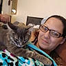 cat, person, portrait, indoors, bedroom, blanket, fluffy, gray_cat, long_hair, whiskers, ears, glasses, selfie, cuddling, relaxed, lying_down, home, furniture, colorful_blanket, pet
