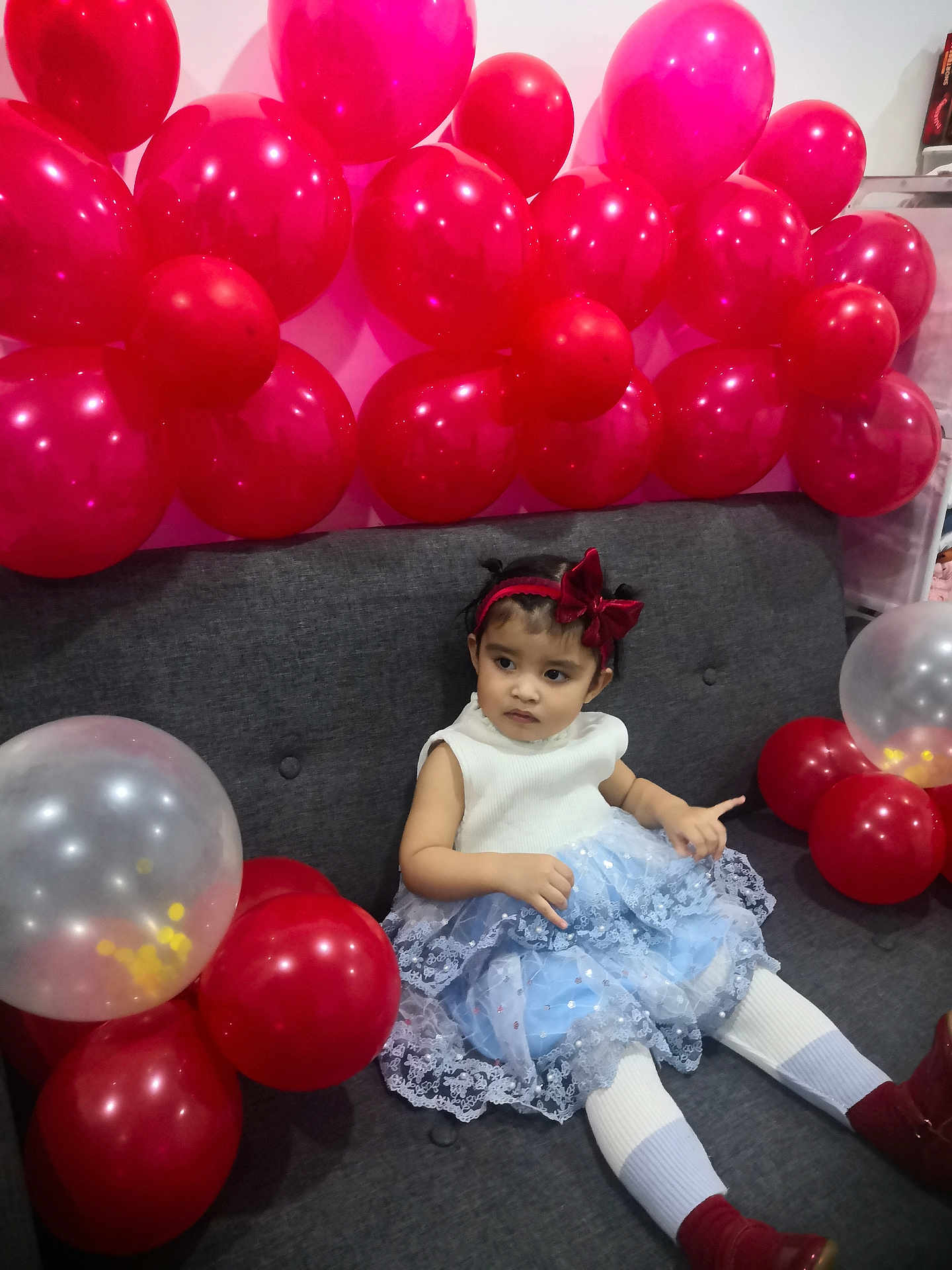 Amber joined the competition — help win amazing prizes! toddler, girl, balloons, red_balloons, transparent_balloons, dress, blue_dress, white_tights, red_boots, headband, red_bow, couch, gray_couch, indoor, celebration, party, child, sitting, curious, festive