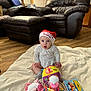 baby, child, christmas_hat, toy, indoor, blanket, floor, wooden_floor, leather_chair, lamp, plush_toy, colorful_toy, sitting, wide_eyes, expression, cute, festive, home, soft_lighting, person