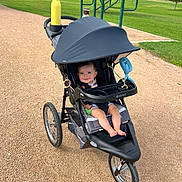 Jack joined the competition — help win amazing prizes! baby, stroller, park, gravel_path, greenery, trees, water_bottle, fan, outdoor, smiling, child, sunny, daytime, grass, wheel, seat, canopy, toy, footwear, person