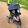 baby, stroller, park, gravel_path, greenery, trees, water_bottle, fan, outdoor, smiling, child, sunny, daytime, grass, wheel, seat, canopy, toy, footwear, person