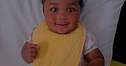 Kennedi is registered to the contest to win money with this photo: baby, child, girl, smiling, bib, headband, pacifier, socks, floral_dress, pillow, yellow_blanket, indoor, cute, happy, toy, face, portrait, sitting, infant, home