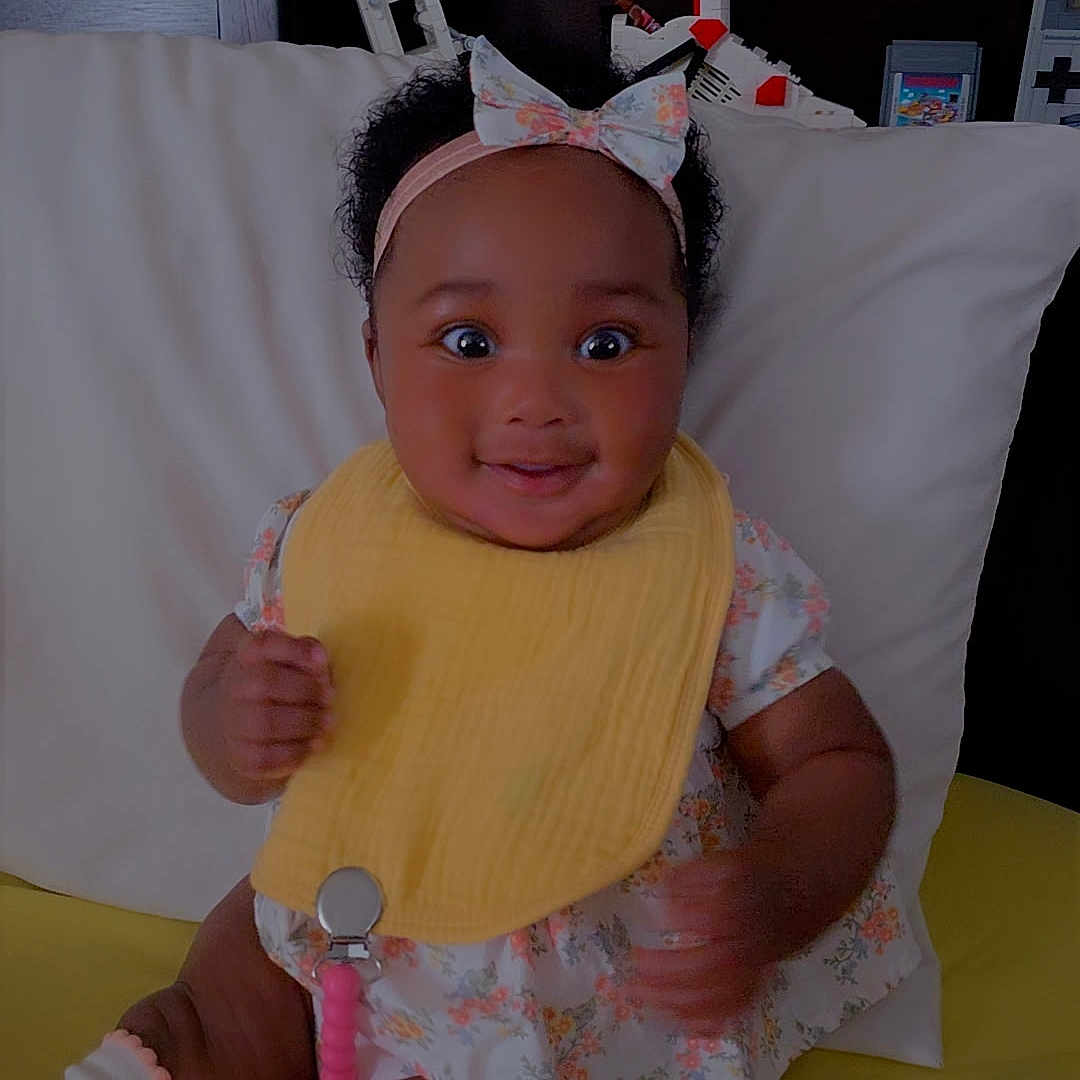 Kennedi is registered to the contest to win money with this photo: baby, child, girl, smiling, bib, headband, pacifier, socks, floral_dress, pillow, yellow_blanket, indoor, cute, happy, toy, face, portrait, sitting, infant, home
