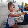 blue_bench, box, casual_clothing, child, concrete_floor, daylight, drink_can, fanta, hair_bun, hand, happy, open_mouth, outdoor, person, picnic_table, playful, sitting, smiling, toddler, white_tank_top