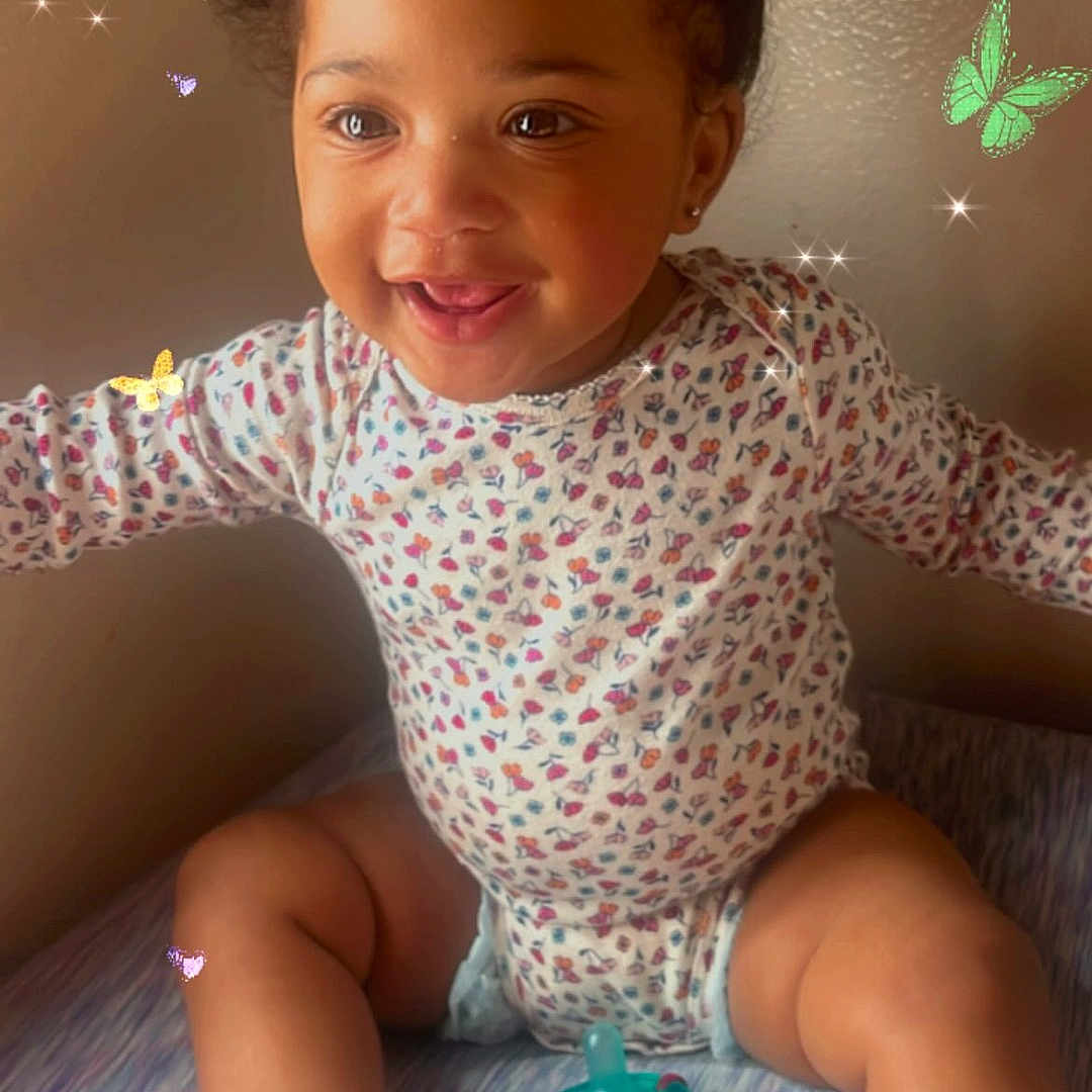 Ahniaya Jones is registered to the contest to win money with this photo: baby, child, smiling, indoor, onesie, socks, pigtails, pacifier, bed, patterned_clothing, cute, happy, skin, wall, butterfly_overlay, digital_effects, infant, young_child, sitting, playful