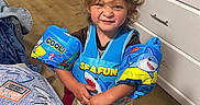 Kairo joined the competition — help win amazing prizes! child, floaties, swim_vest, indoor, wooden_floor, bed, blanket, dresser, clothing, toddler, funny_face, shark, blue, playful, person, floor, furniture, smile, hair, cute