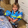 child, floaties, swim_vest, indoor, wooden_floor, bed, blanket, dresser, clothing, toddler, funny_face, shark, blue, playful, person, floor, furniture, smile, hair, cute