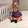 Kairo is registered to the contest to win money with this photo: toddler, child, bed, crib, sandwich, clothing, curly_hair, blue_shirt, spiderman, socks, indoor, bedroom, furniture, person, sitting, food, expression, young_child, blanket, curtains