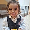 background_blur, child, curly_hair, cute, floor, furniture, happy, holding, indoor, person, playful, portrait, smiling, tile, toddler, toy, vest, white_shirt, yellow, young