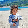 child, curly_hair, grey_sweatshirt, boom_text, outdoor, mountain, lake, sand, nature, casual_clothing, person, sunny_day, blue_sky, greenery, young_child, portrait, expression, standing, discovery, landscape