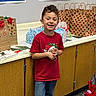Xayden is registered to the contest to win money with this photo: child, boy, smiling, red_shirt, jeans, classroom, gift_bags, holiday, decorations, cabinet, carpet, poster, wall, paper_bags, red_bows, hand_clasp, standing, happy, indoor, education