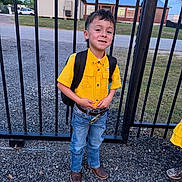 Xayden joined the competition — help win amazing prizes! child, boy, yellow_shirt, blue_jeans, backpack, cowboy_boots, fence, gravel, outdoor, daylight, smiling, standing, casual_clothing, short_hair, portrait, school_ready, metal_bars, building, grass, road
