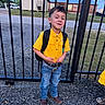 backpack, blue_jeans, boy, building, casual_clothing, child, cowboy_boots, daylight, fence, grass, gravel, metal_bars, outdoor, portrait, road, school_ready, short_hair, smiling, standing, yellow_shirt