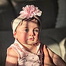 child, baby, toddler, portrait, face, eyes, sitting, pink_clothing, floral_headband, hair_accessory, dress, patterned_clothing, earring, indoor, warm_lighting, sunlight, window_light, closeup, person, photography