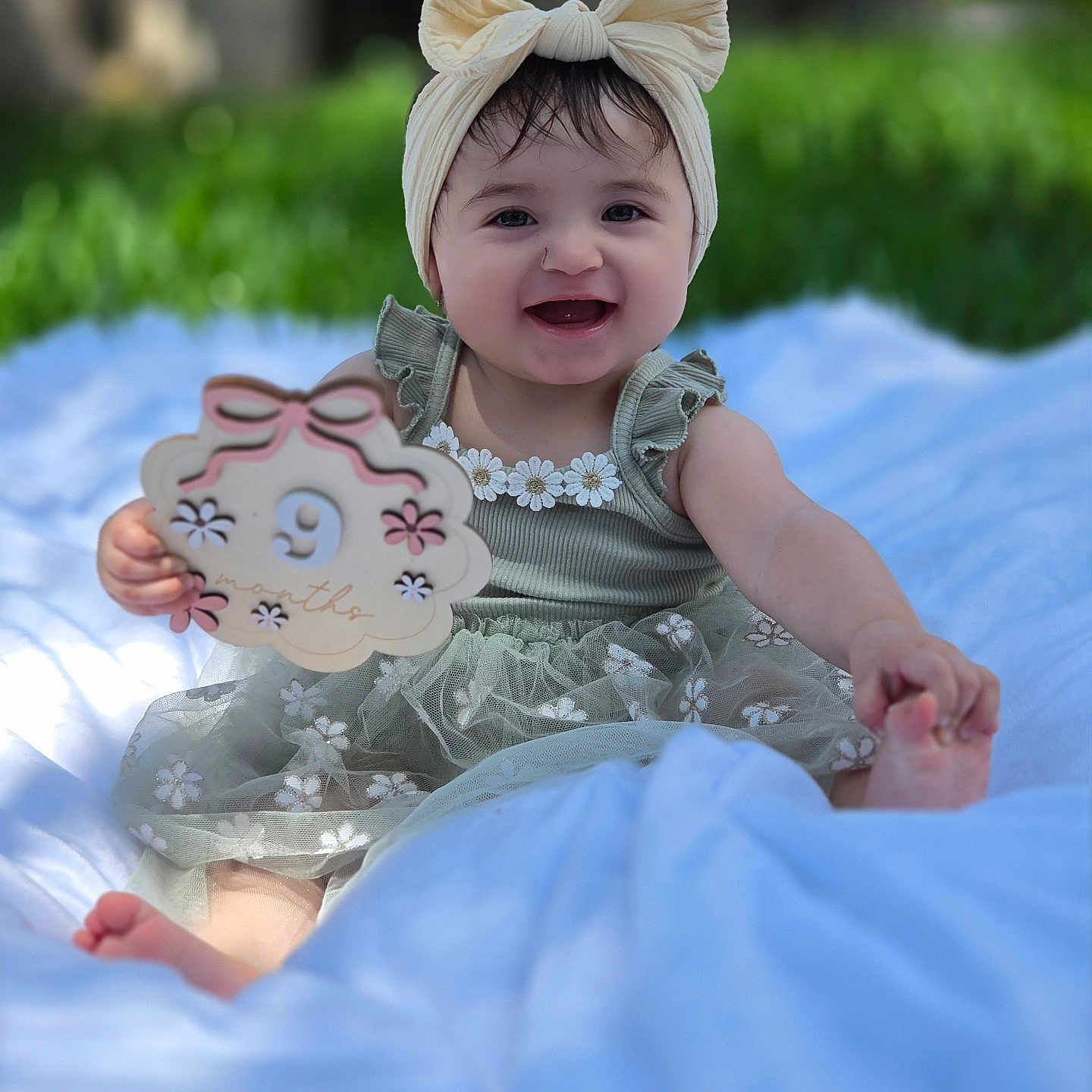 Danelly Anais is registered to the contest to win money with this photo: baby, child, smiling, portrait, outdoor, garden, blanket, blue_blanket, headband, bow, dress, floral_dress, sitting, happy, infant, toy_sign, birthday, greenery, bokeh, feet