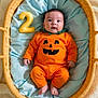 baby, infant, pumpkin_costume, orange_clothing, wicker_basket, blue_cushion, number_two, plush_toy, cute, holiday, halloween, child, portrait, indoors, soft_lighting, smiling_face, cozy, comfort, sitting, celebration