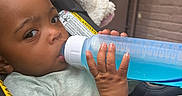 Damarionna joined the competition — help win amazing prizes! baby, child, infant, car_seat, bottle, water, drinking, portrait, face, hand, eyes, smiling, indoors, vehicle, safety_harness, gray_clothing, blue_bottle, close_up, seated, transportation