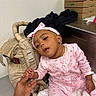 baby, child, infant, portrait, pink_clothing, floral_pajamas, headband, bow, smiling, sitting, hand, adult_hand, blanket, baby_carrier, indoor, office, cardboard_boxes, subway_boxes, desk, closeup