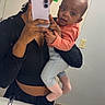 woman, baby, mirror, selfie, bathroom, sink, phone, clothing, headband, floor, tile, light_switch, holding, portrait, smiling, person, child, casual, indoor, reflection