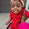 baby, child, tongue_out, red_clothing, pink_bow, headband, hand, outside, brick_wall, door, playful, portrait, infant, cute, smiling, person, holding, young_child, casual, expression