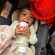 Damarionna is registered to the contest to win money with this photo: baby, infant, car_seat, blanket, red_blanket, milk_bottle, white_object, clothing, cute, child, indoors, portrait, hands, face, happy, feeding, soft_texture, warm, seated, curious