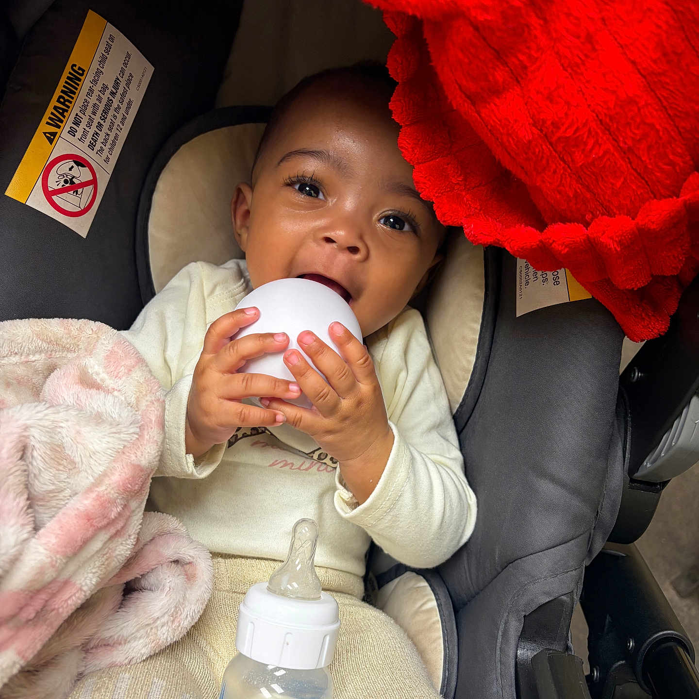 Damarionna is registered to the contest to win money with this photo: baby, infant, car_seat, blanket, red_blanket, milk_bottle, white_object, clothing, cute, child, indoors, portrait, hands, face, happy, feeding, soft_texture, warm, seated, curious