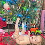 baby, blanket, bokeh_lights, child, christmas_lights, christmas_tree, curly_hair, diaper, festive, floor, holiday_decor, indoor, ornament, pine_branches, playful, red_green_pattern, smile, toddler, toy_truck, tree_skirt