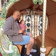 Emmie a rejoint le concours — aidez-le/la à gagner de superbes lots ! man, baby, carousel, amusement_park, ride, ornate, decorative, seat, casual_clothing, shorts, jacket, sneakers, child, parent, outdoor, daylight, family, fun, leisure, people