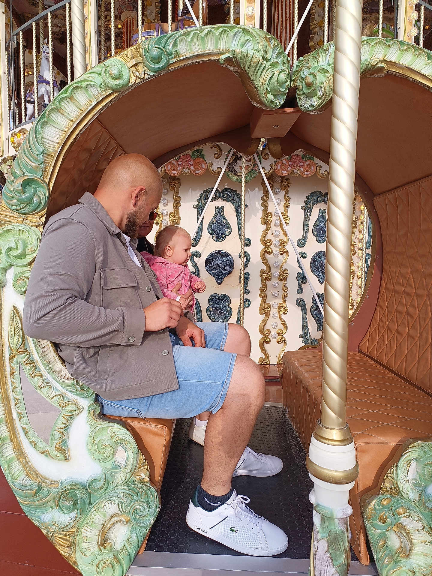 Emmie a rejoint le concours — aidez-le/la à gagner de superbes lots ! man, baby, carousel, amusement_park, ride, ornate, decorative, seat, casual_clothing, shorts, jacket, sneakers, child, parent, outdoor, daylight, family, fun, leisure, people