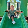 active, casual_clothing, child, daylight, fun, green_slide, happy, outdoor, person, play, playground, recreation, shorts, slide, smiling, sneakers, summer, sunny, toddler, tree