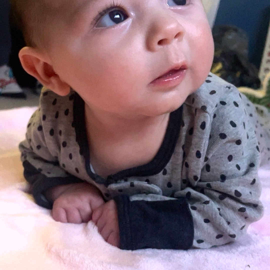 Liora is registered to the contest to win money with this photo: baby, infant, child, face, portrait, curious, looking_up, eyes, smiling, indoors, blanket, pink_blanket, polka_dot, clothing, sitting, lying_down, home, window, soft_lighting, close_up